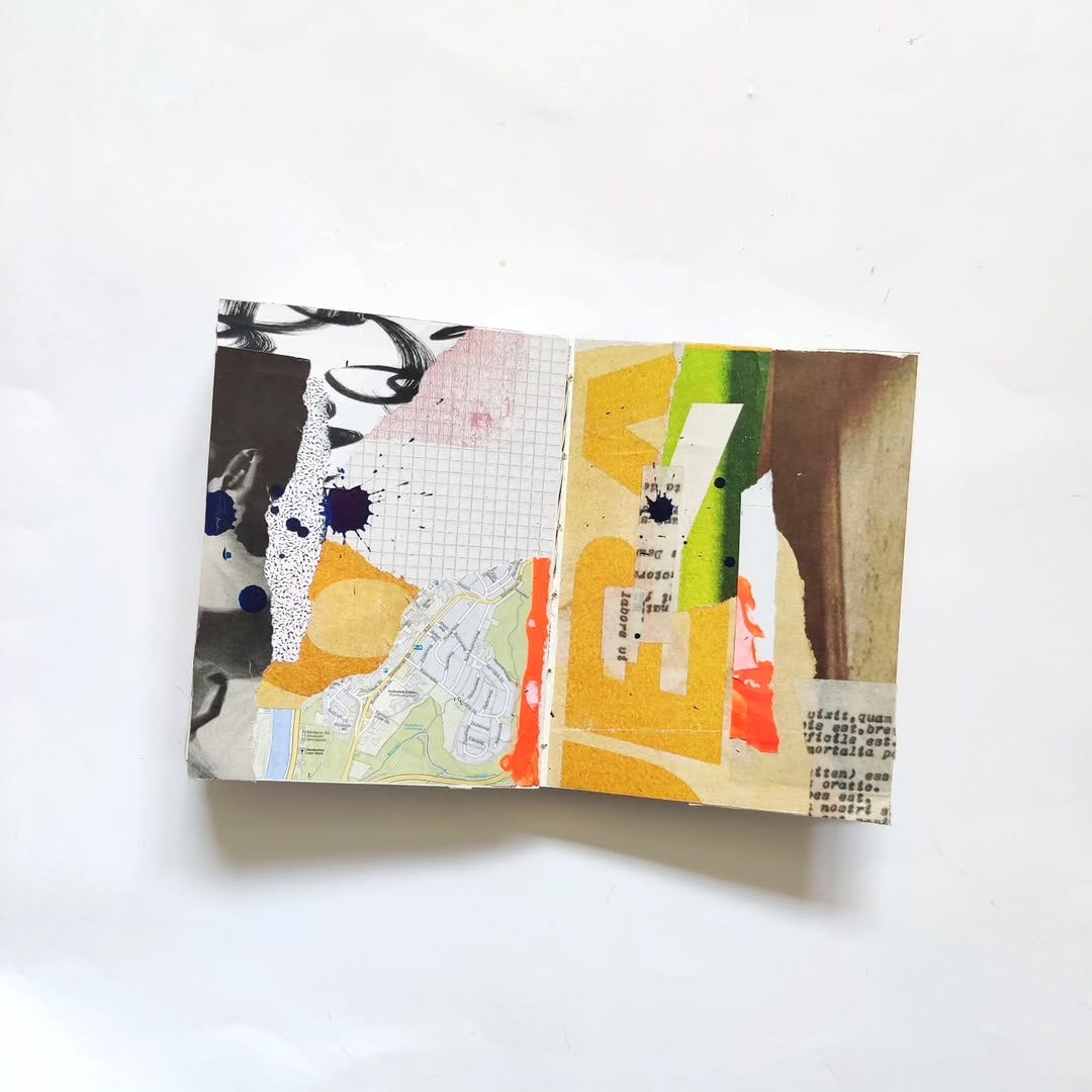 Open mixed media art journal. A layer of collage — mostly neutral tones, with some greens, yellows, and oranges — is topped with a few splatters of black ink.