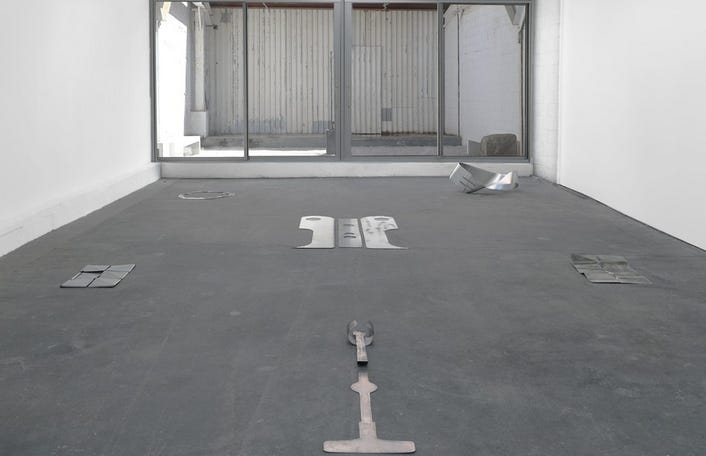 Metal forms, ranging from the size of a laptop to a guitar, that appear flatly on the floor of a white-walled gallery. A backyard sculpture garden with rocks exists in the background. Metal forms, ranging from the size of a laptop to a guitar, that appear flatly on the floor of a white-walled gallery. A backyard sculpture garden with rocks exists in the background.