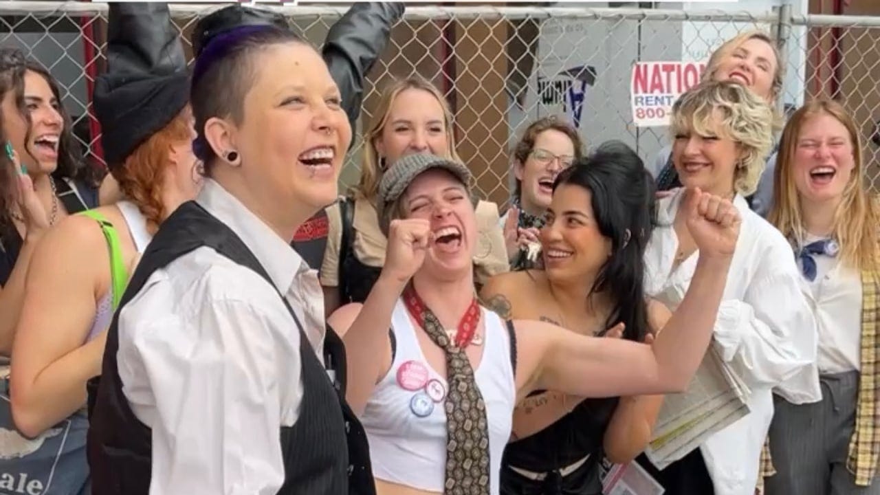 Women stand next to a chain-link fence smiling, laughing and celebrating.