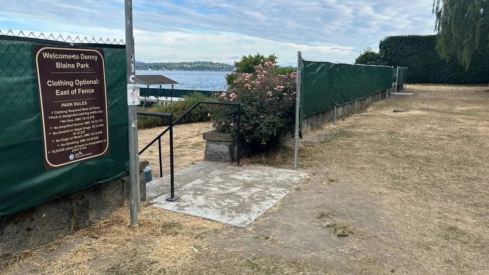 The entrance to Denny Blaine Park showing a brown sign reading "Welcome to Denny Blaine Park — Clothing Optional: East of Fence" with park rules listed below, next to a chain-link fence wrapped in green privacy mesh, with Lake Washington visible in the background.