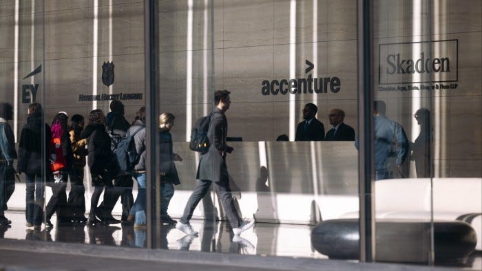 People walk past a reception desk with the Accenture logo in a lobby, with other company logos visible on the wall.