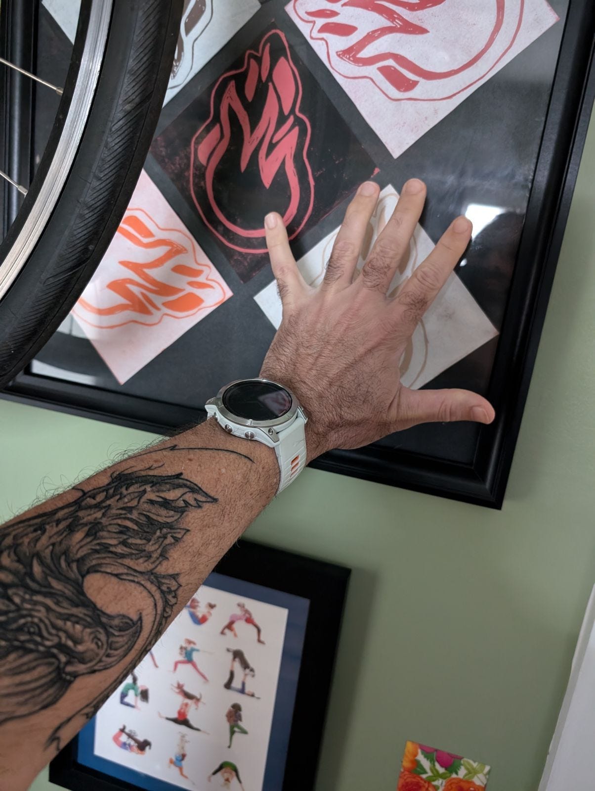 A close-up photo of the author’s forearm and hand. He is holding his hand up to a block-printed art with a black background. Hot-colored fire illustrations printed on white diamonds, X4, with one in the middle using negative space- red on black. His hand is spread over the bottom-right flame to visualize its placement for the tattoo. A close-up photo of the author’s forearm and hand. He is holding his hand up to a block-printed art with a black background. Hot-colored fire illustrations printed on white diamonds, X4, with one in the middle using negative space- red on black. His hand is spread over the bottom-right flame to visualize its placement for the tattoo.