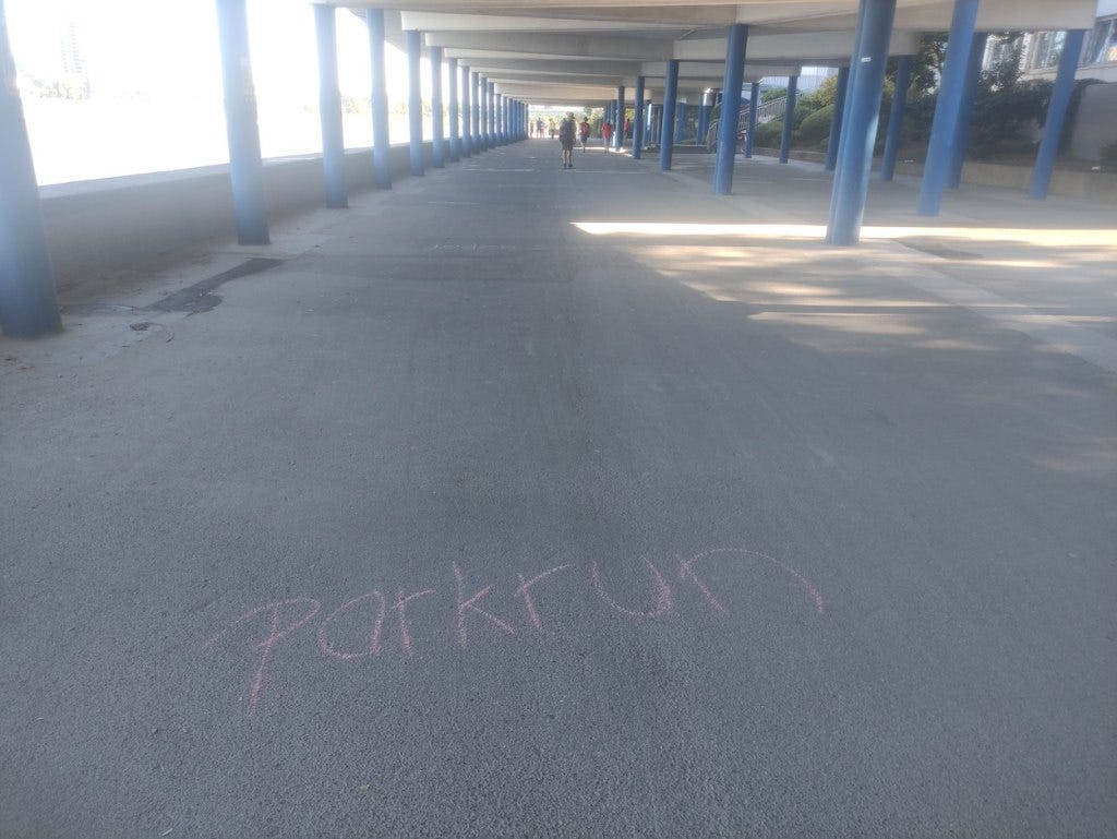 The word parkrun is chalked onto the tarmac, with further words explaining the concept just out of sight, further along the route. Blue pillars hold a concrete roof over this part of the riverside walkway