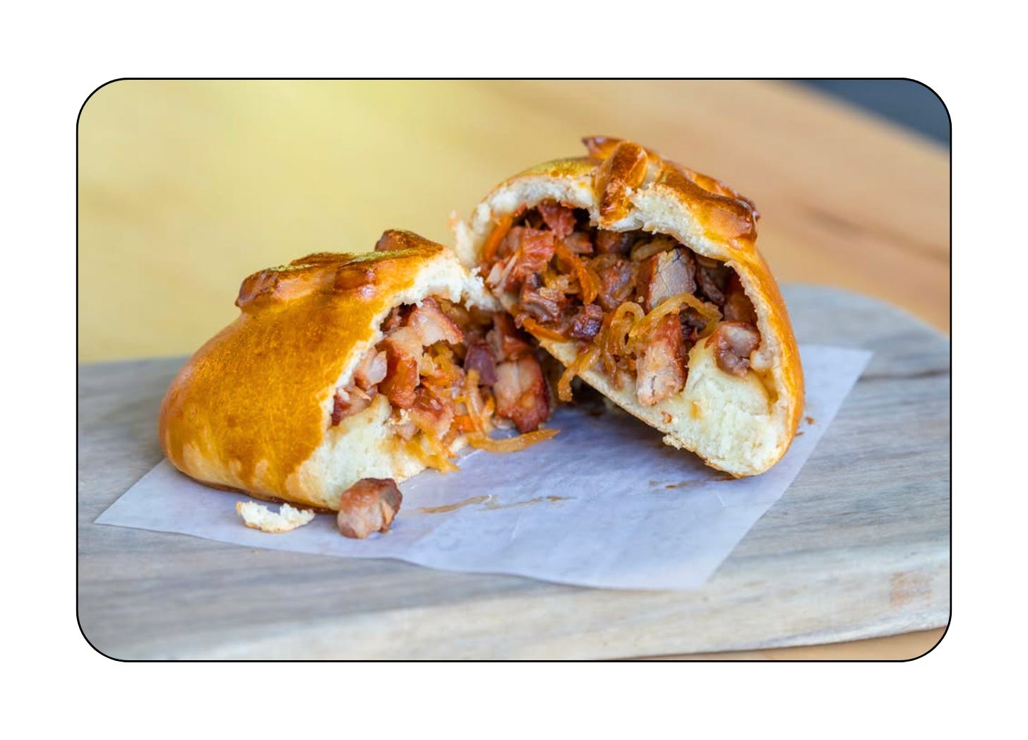 A piroshki split in half revealing a pork liempo filling, resting on parchment paper on a wooden board.