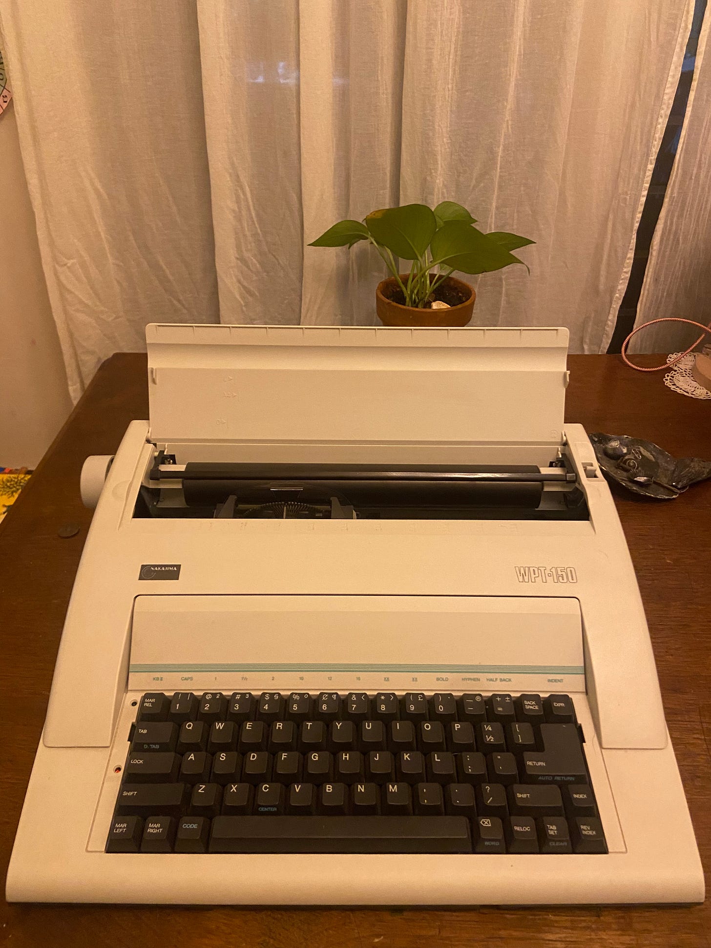 White Nakajima electronic typewriter on a wooden desk with a small pothos plant behind it. White Nakajima electronic typewriter on a wooden desk with a small pothos plant behind it.