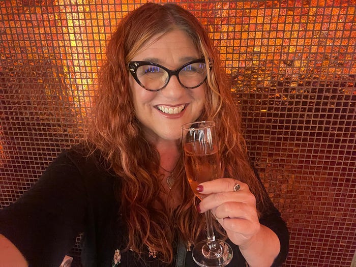 The author holding a glass of sparkling rose in front of a tiled wall that matches her hair. The author holding a glass of sparkling rose in front of a tiled wall that matches her hair.