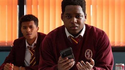 Two boys sit in a classroom facing forward and one, holding a phone in his hand, looks very concerned about something
