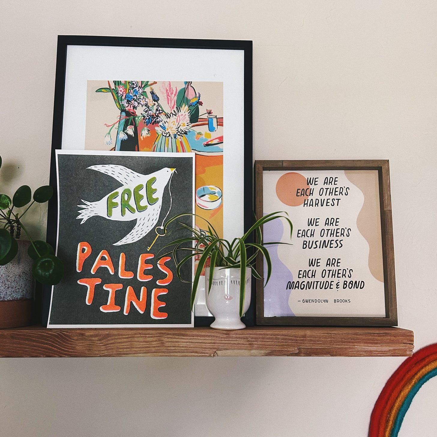 Image ID: Photo of a wooden shelf with plants and art prints on it. One says Free Palestine with a dove holding a key, and the other is a quote by Gwendolyn Brooks: We are each other's harvest. We are each other's business. We are each other's magnitude and bond.
