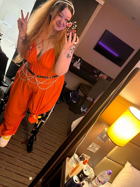 photo 1: A person seated in a wheelchair, smiling and holding up an iPad showing their career journey. They’re in a cosy room with yellow walls, autumn leaf decorations, and plants in the background.  photo 2: A mirror selfie in a hotel room. The person is wearing a bright orange jumpsuit with chain accessories, long orange hair, and giving a peace sign while seated in a wheelchair.  photo 3: A person sitting on a bed wearing a striped dress and a decorative tiara, smiling warmly. The room has colourful bedding, wall hangings, and soft string lights.  photo 4: A person in a wheelchair reaching through a fence to pet a Highland cow in a field. The scene is outdoors with fallen autumn leaves and trees with golden foliage.  photo 5: A mirror selfie taken in an accessible bathroom. The person is wearing sunflower-patterned clothing and a bright yellow top, smiling while seated in a wheelchair.