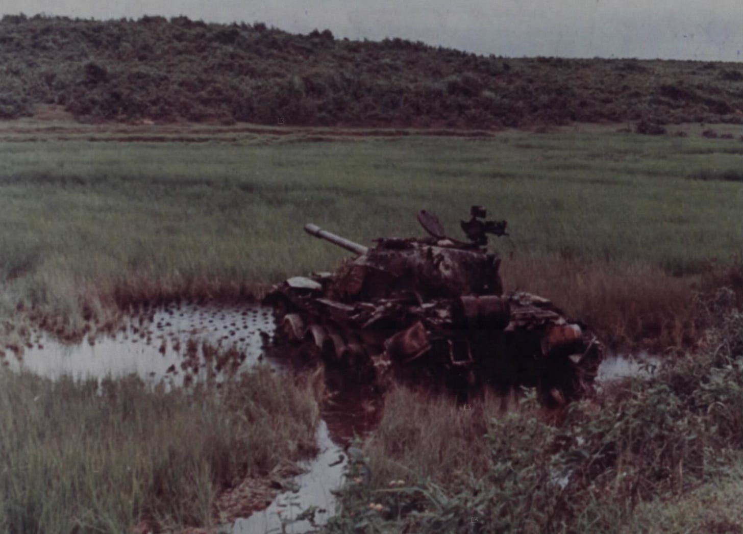 A wrecked tank sitting tilted in a flooded rice paddy, with grass in front and low hills behind. A wrecked tank sitting tilted in a flooded rice paddy, with grass in front and low hills behind.