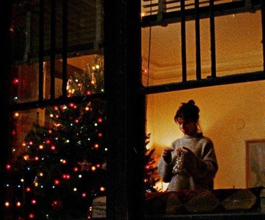 This may contain: a woman standing in front of a christmas tree looking out the window at her cell phone