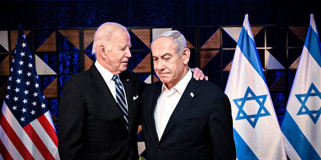 Joe Biden in a dark suit rests his hand on Netanyahu's shoulder as they stand together in front of U.S. and Israeli flags