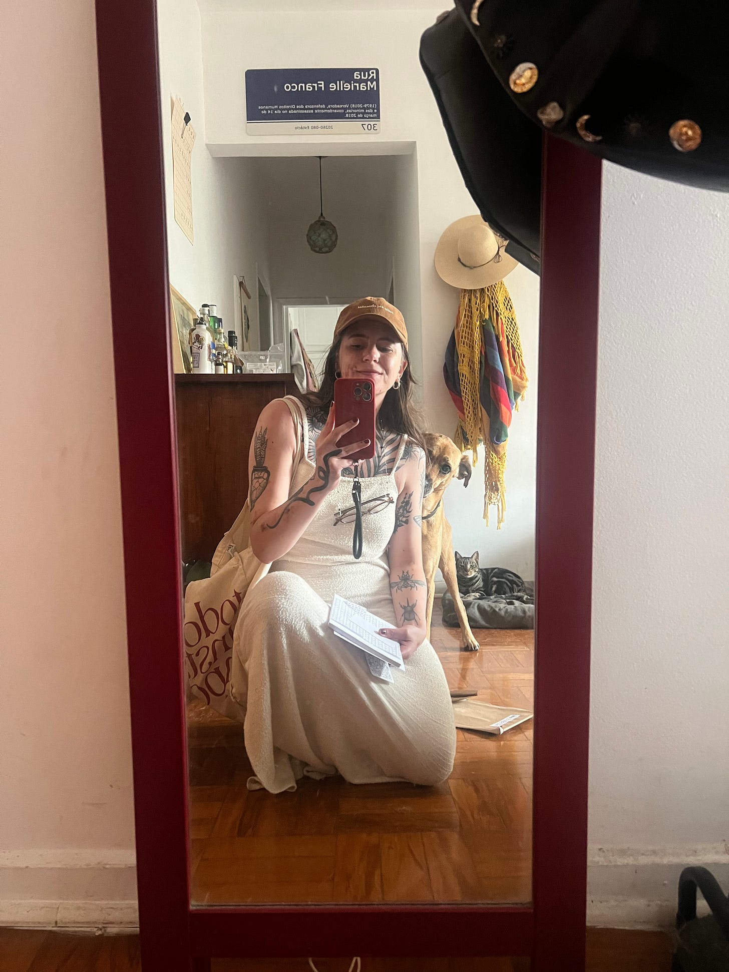 Foto no espelho de casa, estou agachada no chão, usando um vestido branco longo, um boné marrom, e logo atrás de mim tem o Batista meio se escondendo. Mais ao fundo, o gato Fortunato está deitado na cama do cão