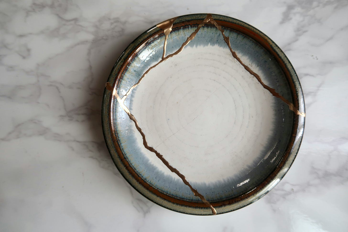 A ceramic plate with a blue-grey glazed rim and cracks repaired with gold kintsugi seams, sitting on a white marble surface.