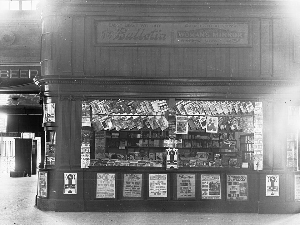 A black and white photograph of the side of a storefront. There are two large open windows on the side with magazines or newspapers hanging from the top of the window. Below the window there are advertising displays of what looks like newspaper front pages.