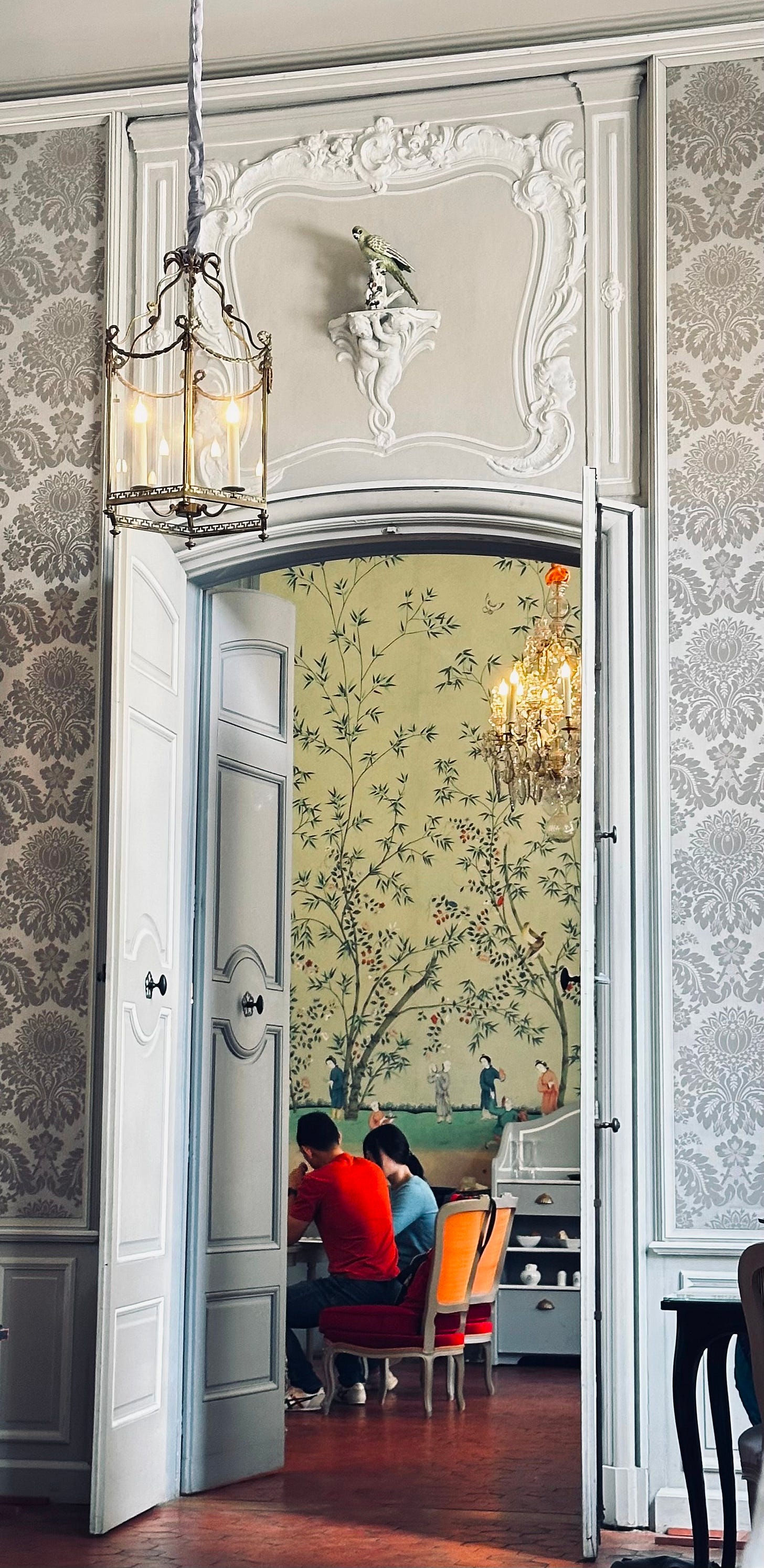 Open white double doors inside an elegant European interior, revealing a softly lit room beyond where two people sit at a table beneath a chandelier and painted wall mural — a quiet moment of entering and belonging. Open white double doors inside an elegant European interior, revealing a softly lit room beyond where two people sit at a table beneath a chandelier and painted wall mural — a quiet moment of entering and belonging.