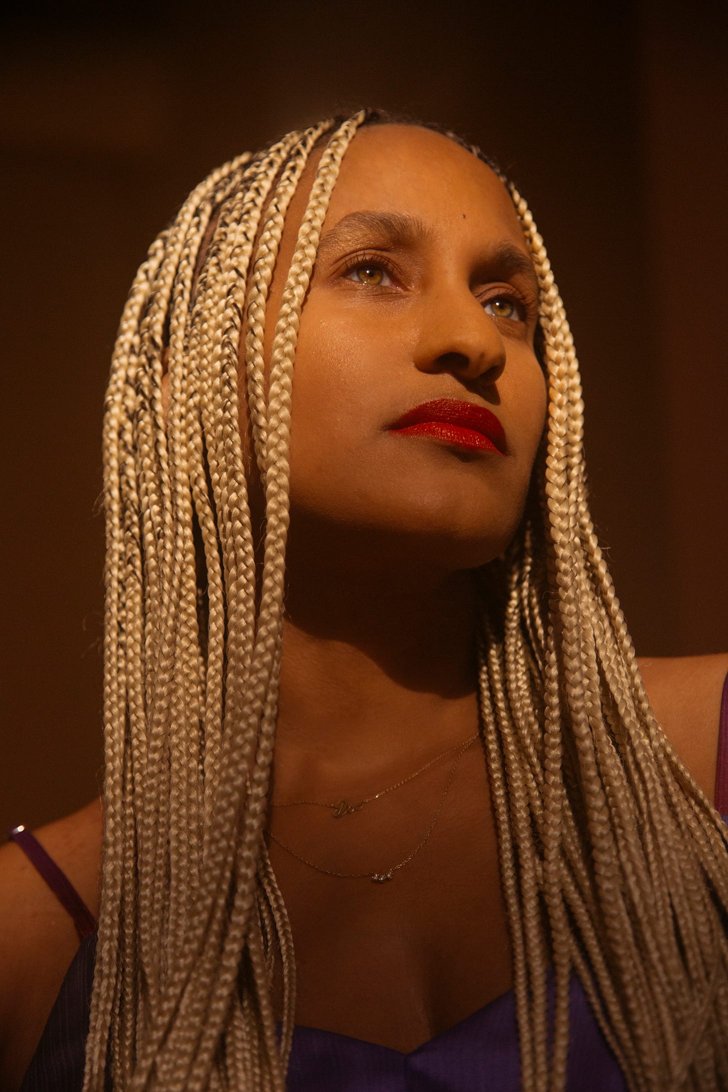 Dr. Akilah Cadet with long, light blonde braids and red lipstick gazes upward in warm, dramatic lighting. She wears a purple top and layered gold necklaces. Dr. Akilah Cadet with long, light blonde braids and red lipstick gazes upward in warm, dramatic lighting. She wears a purple top and layered gold necklaces.