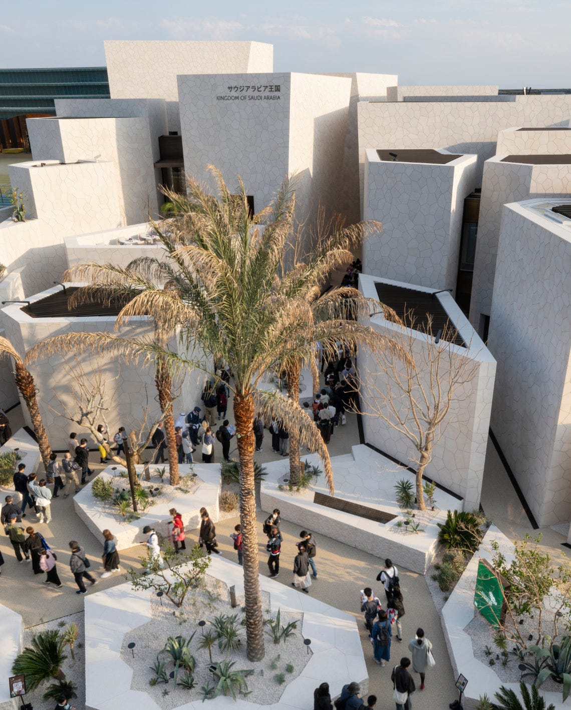 Visitors walk through a courtyard with palm trees and modern white stone buildings at the Saudi Arabia Pavilion, Expo 2025.