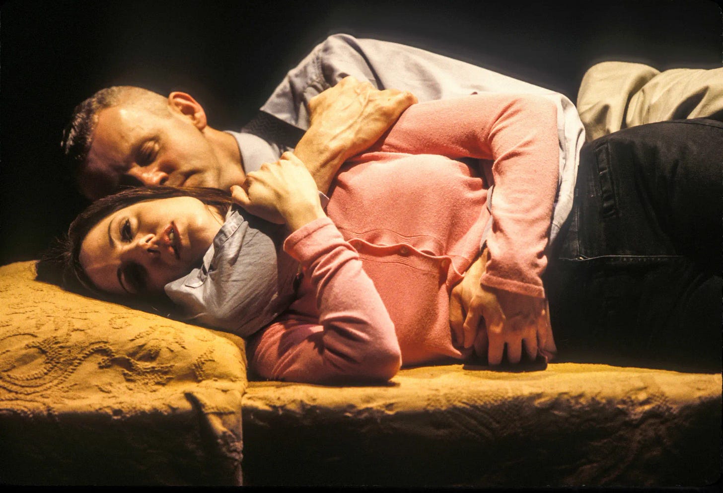 A white man hugs a white woman from behind. Both are clothed and lying on a bed.