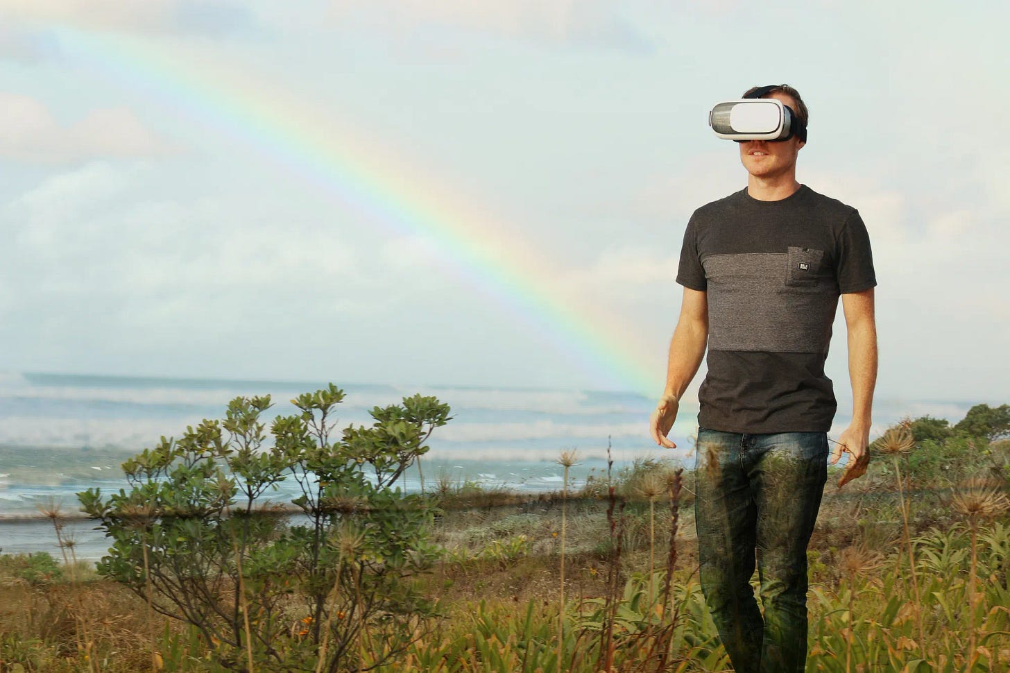 Man in field with rainbow and ocean in background walking with virtual reality headset over his eyes.
