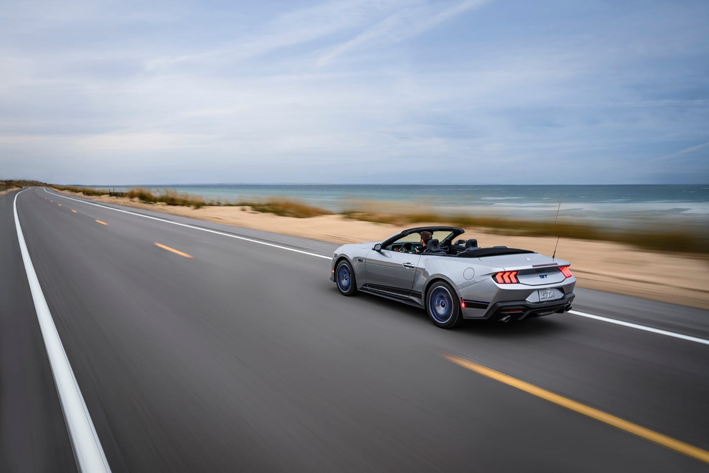 A silver 2024 Ford Mustang GT California Special convertible drives on a two-lane highway next to an ocean and beach, with the top down.