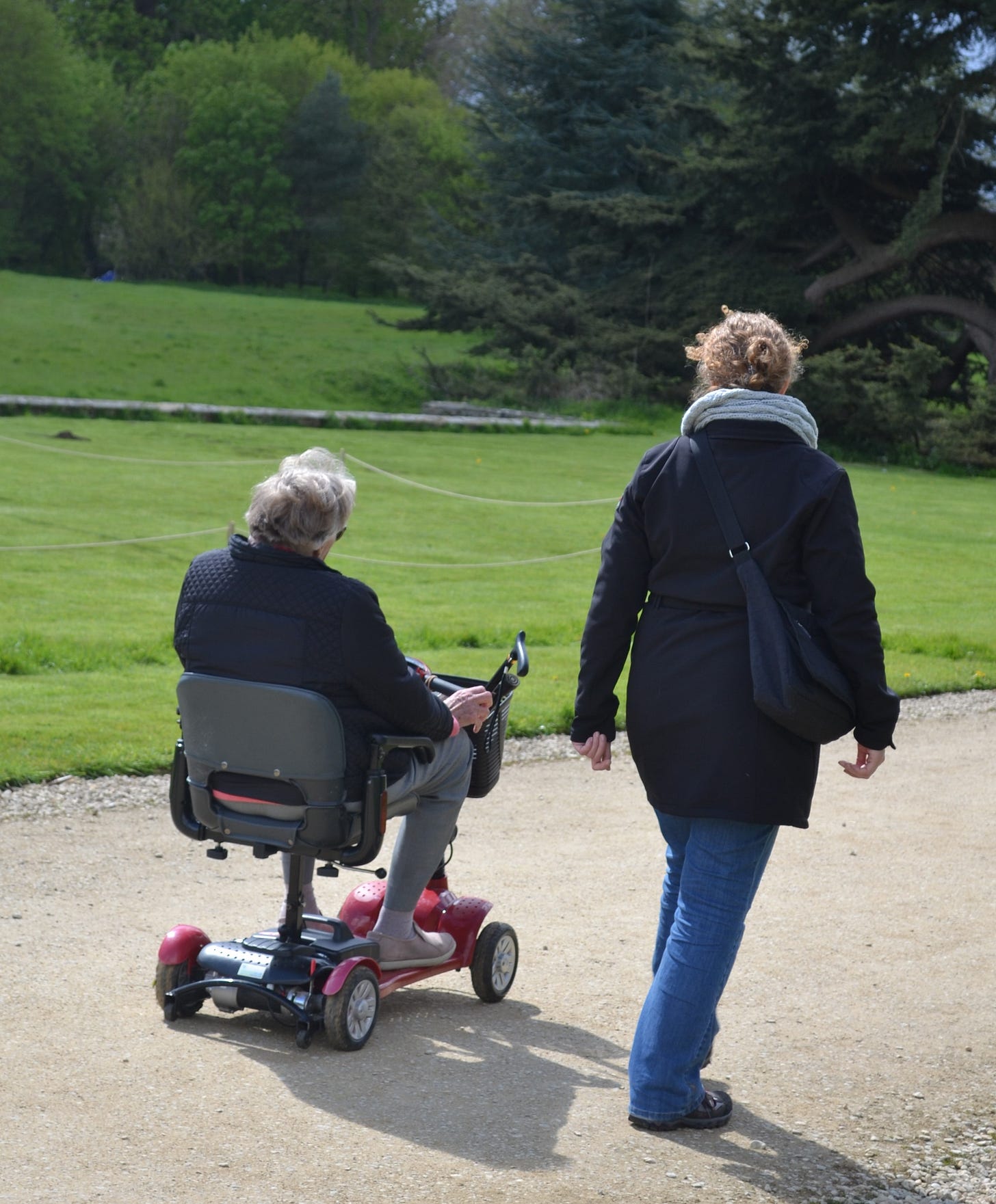 My mum, in her 80s and on a mobility scooter, and me, heading along a compacted gravel path, surrounded by trees and beautifully-mown lawns.