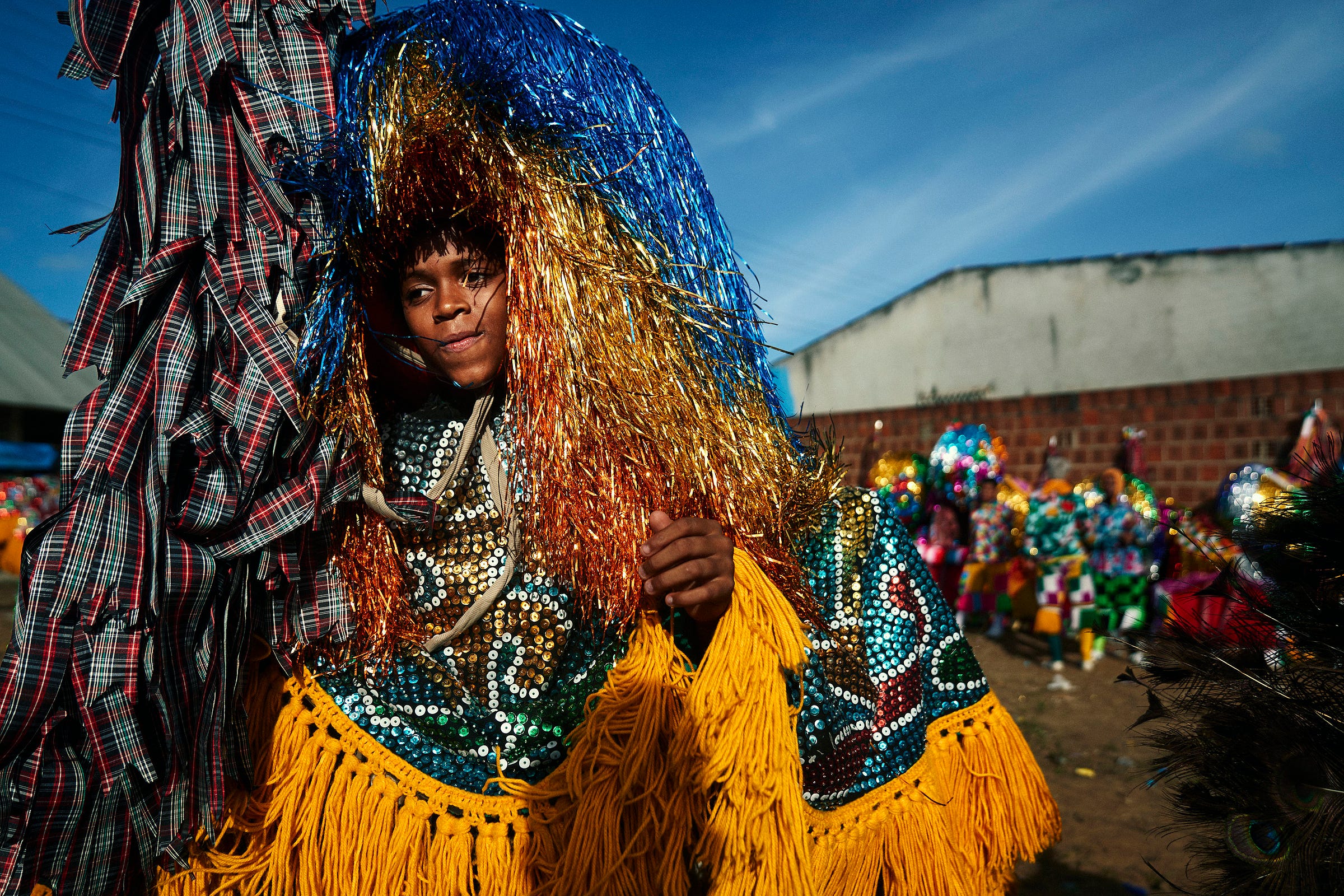A person wearing a vibrant costume adorned with yellow fringe, blue and orange tinsel, and sequins stands outdoors at a festival, with colorful decorations and blue sky in the background.