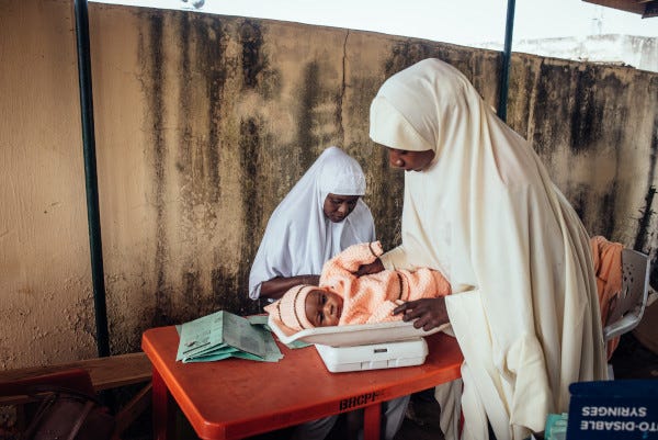 A mother and child wait at a clinic in Jigawa while a Routine Immunization Provider prepares paperwork before administering the infant's routine childhood immunizations. A mother and child wait at a clinic in Jigawa while a Routine Immunization Provider prepares paperwork before administering the infant's routine childhood immunizations.