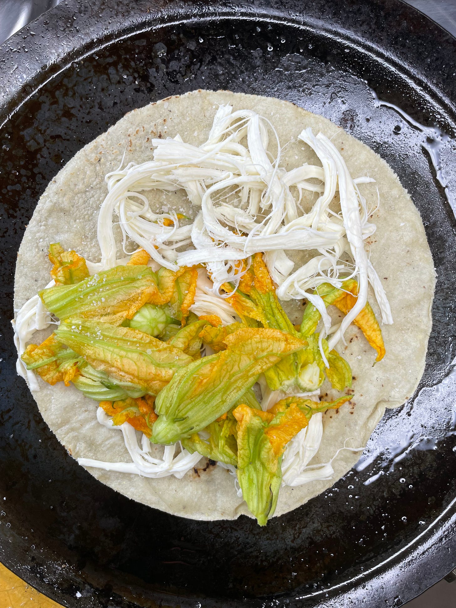 A tortilla on a greased comal covered with cheese and squash blossoms.