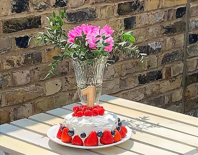 A small white-frosted cake topped with strawberries, blueberries, raspberries, and a gold number “1” candle sits on a white outdoor table. Behind it, a glass vase holds a bouquet of pink flowers and greenery. A sunlit brick wall forms the background.