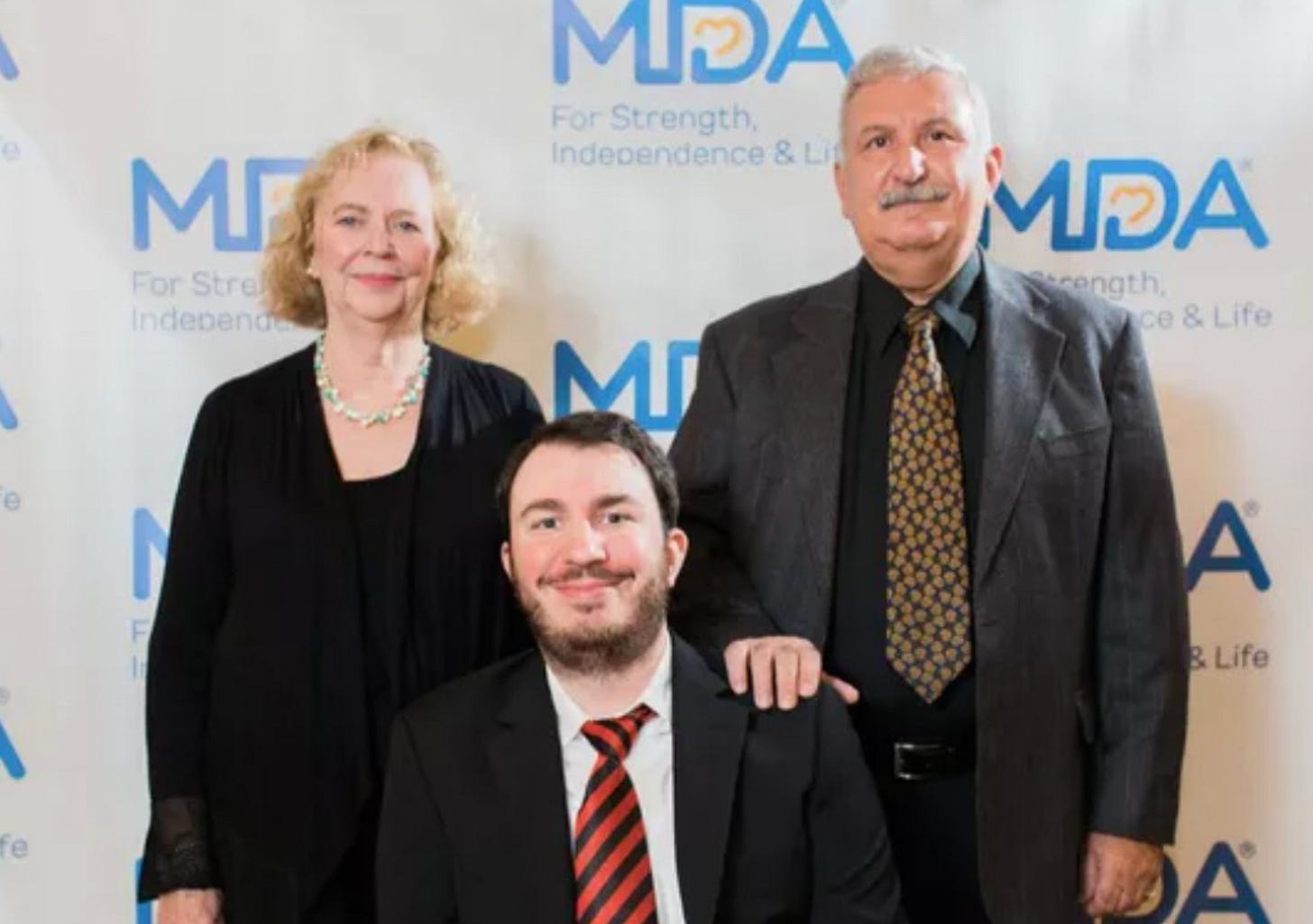 Chris, a young man in a wheelchair wearing a black suit and red striped tie, poses with his parents at a Muscular Dystrophy Association event. His mother stands to the left wearing black with a green necklace, and his father stands to the right in a dark suit with a patterned tie, his hand resting supportively on Chris's shoulder. The MDA logo appears repeatedly in the background.