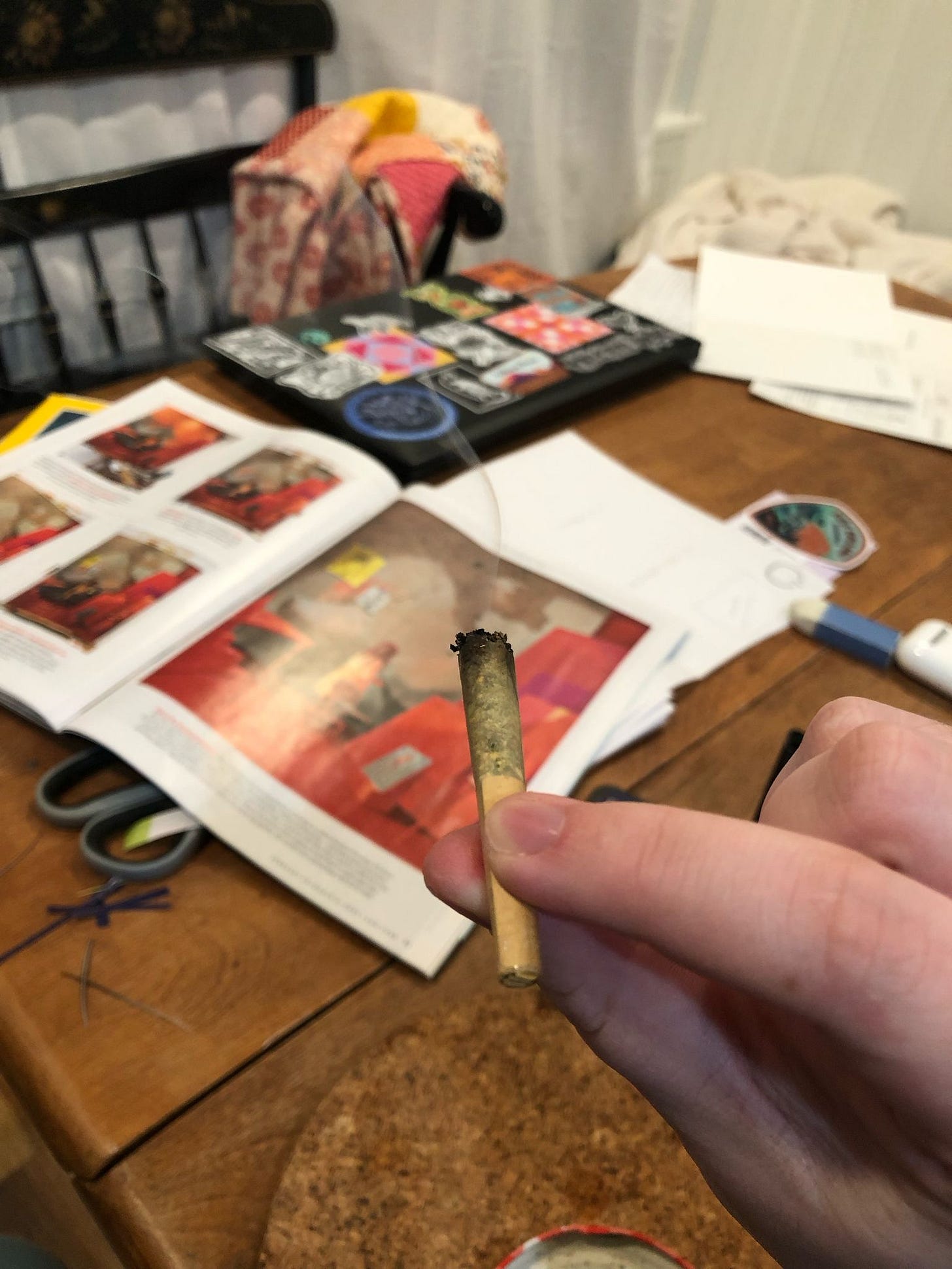 A hand holding a lit joint in the foreground, with a table covered in collage supplies in the background