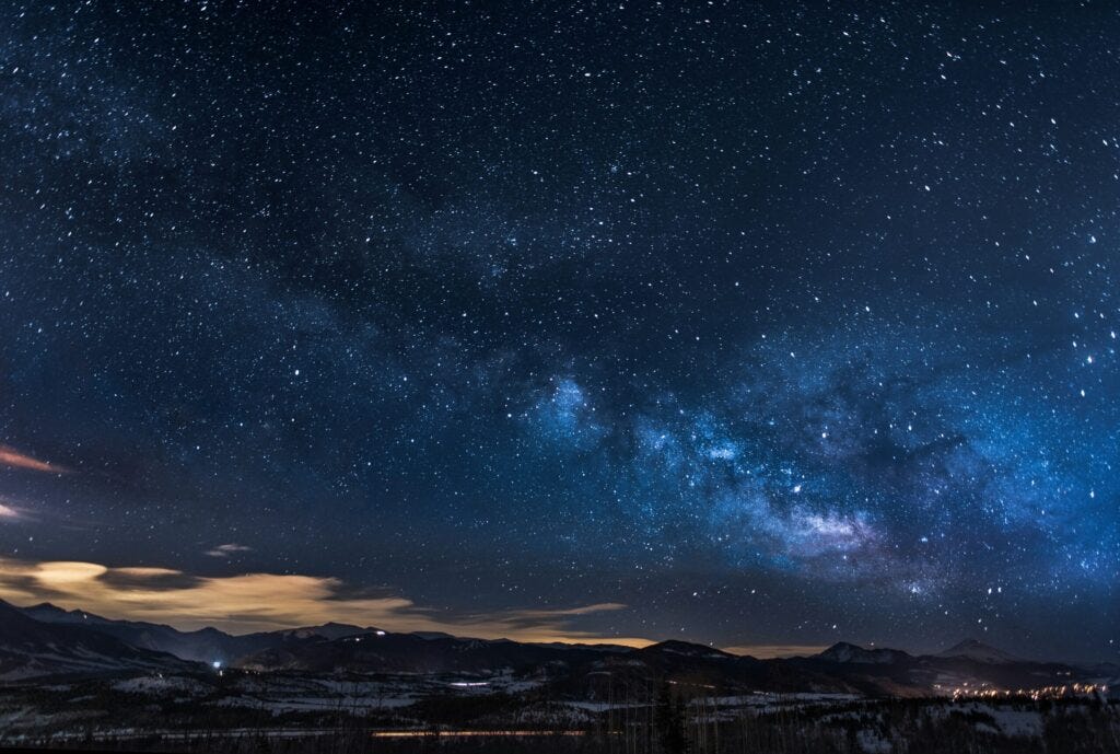 A dark night sky with stars above, clouds and hills below.