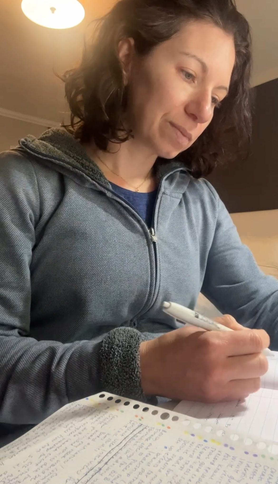 A woman with dark hair wearing a blue hoodie writing in her notebook