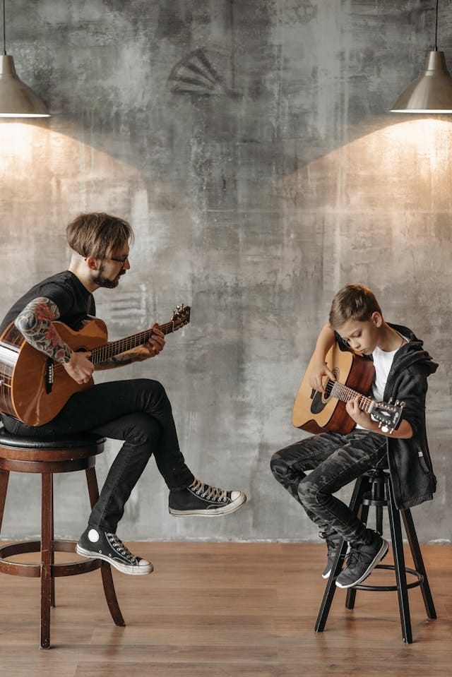 Two men sitting on stools, playing a classical guitar. The older one is teaching the younger one, a pre-teen, how to play guitar.