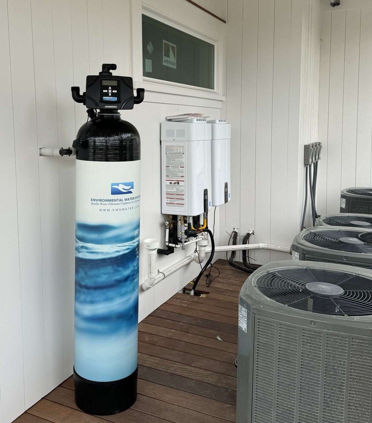 A tall whole-house water filtration tank with a black control head standing against white shiplap on a covered porch, with two tankless water heaters mounted on the wall behind it and outdoor AC condensers to the right. A tall whole-house water filtration tank with a black control head standing against white shiplap on a covered porch, with two tankless water heaters mounted on the wall behind it and outdoor AC condensers to the right.