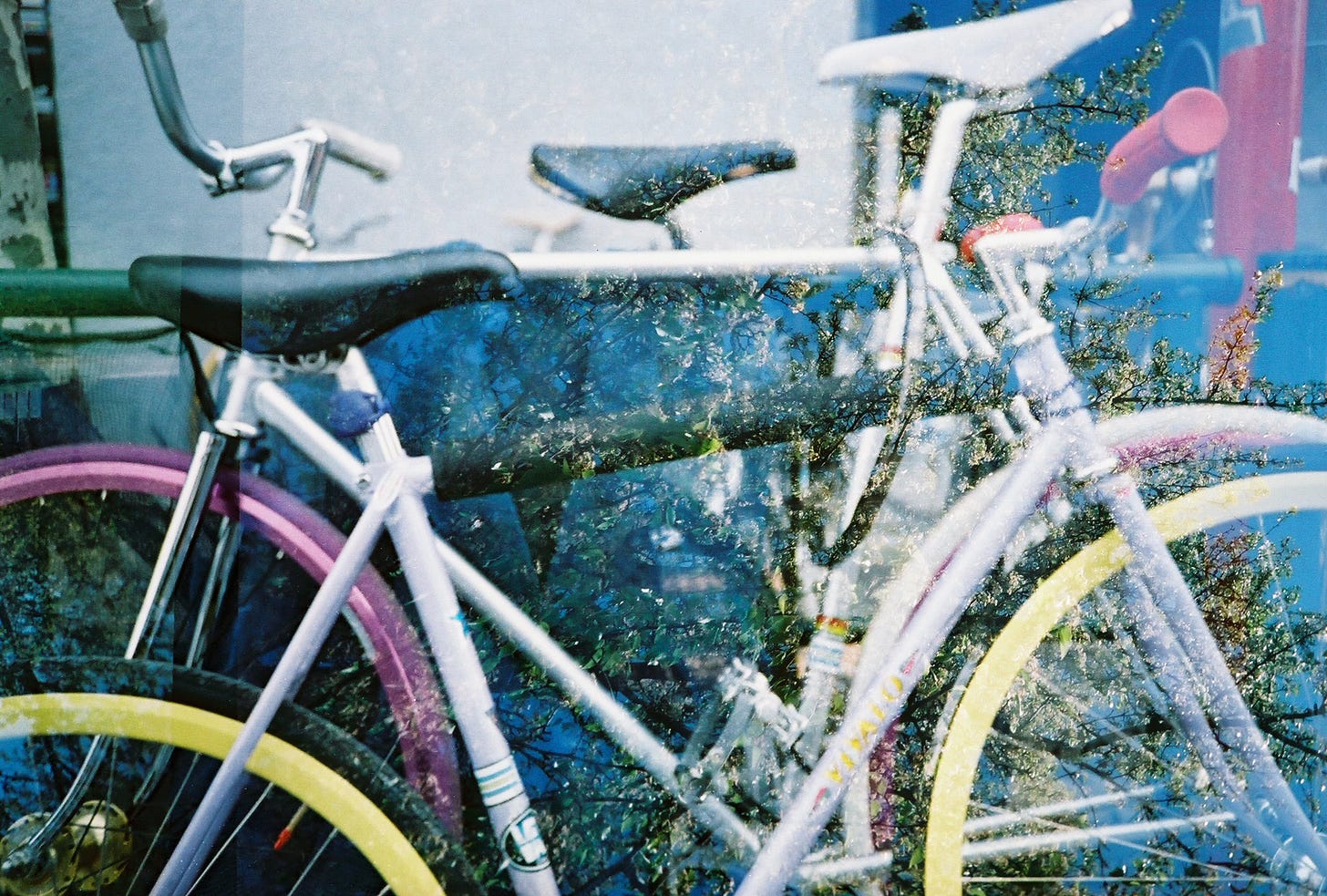 A double exposed film photo shows a close-up image of multi-colored bikes against a photograph of green trees and a blue sky.