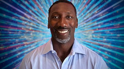 Jimmy Floyd Hasselbaink smiles for the camera wearing a light blue shirt, set against a glittering blue background