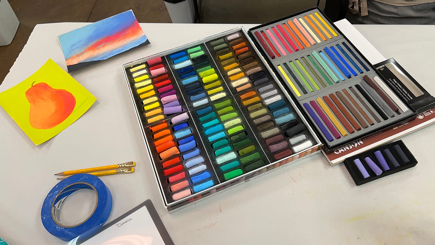 picture of a table with soft pastels in boxes with a painting of a coral colored pear on a neon yellow green background and an attemopt at painting a sunset over mountains