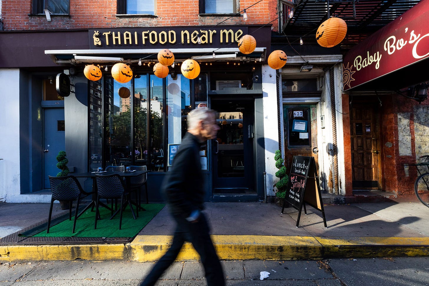 Thai Food Near Me restaurant seen from the street. A person is walking by, and there are Halloween decorations hanging above the sign.