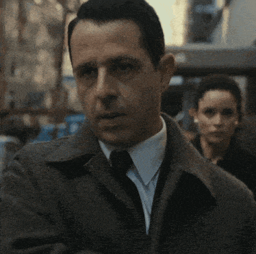 a man in a suit and tie with a woman in the background a man in a suit and tie with a woman in the background