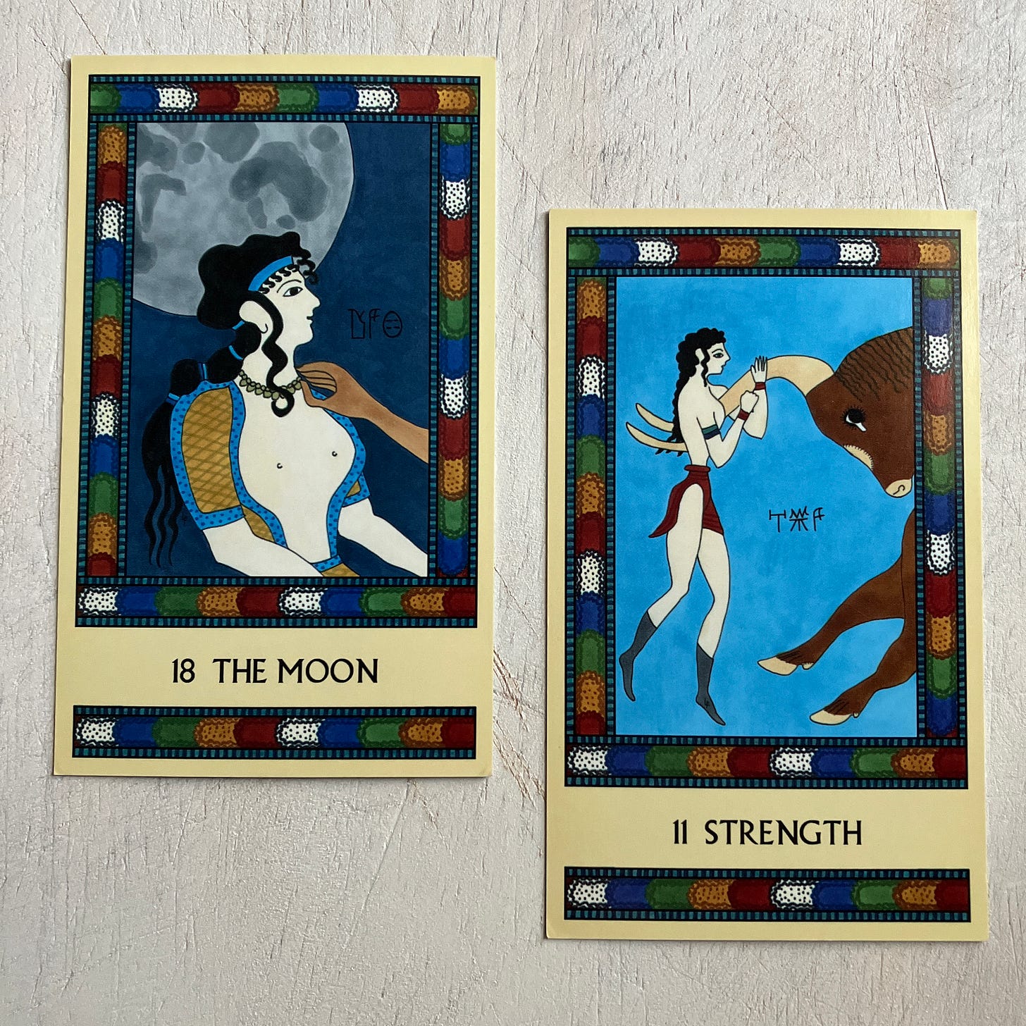 Two Minoan Tarot cards on an off-white painted wood surface. The Moon shows a white-skinned Minoan woman facing to her left as a brown-skinned hand adjusts her necklace while a full moon hangs in the sky above. Strength shows a loincloth-clad white-skinned female bull leaper hanging on the long, curving horns of a brown bull in preparation for the leap. 