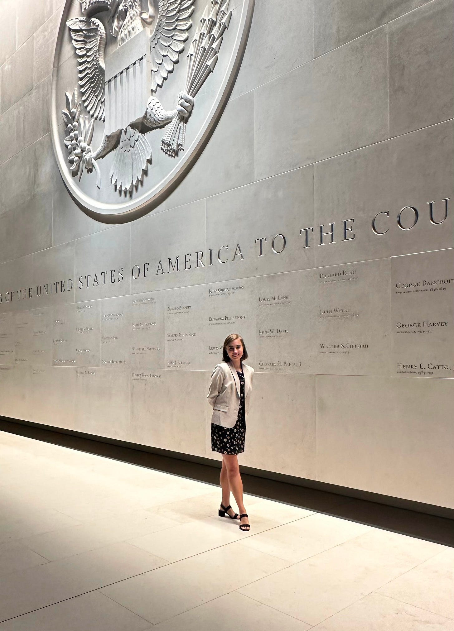 A woman wearing a tan blazer and black dress standing in front of a grey stone wall