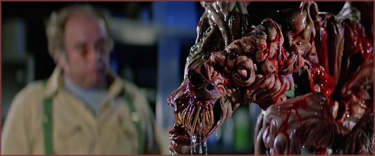Film frame from The Thing (1982): Blair (Wilford Brimley) out of focus in the background, while the remains of the Dog-Thing creature fill the foreground in sharp detail — a mass of foam latex flesh, teeth, and biological-looking fluid constructed by Rob Bottin's effects team.