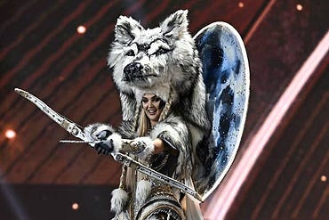 Person in an elaborate costume with a large wolf headpiece and shield, holding a bow; appears to be on stage