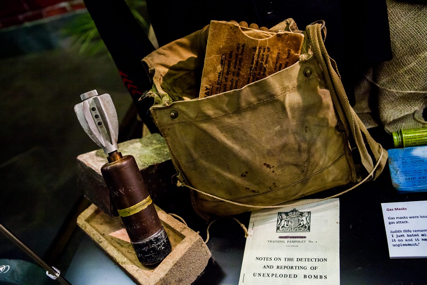 A small replica bomb next to a canvas satchel with training leaflets sticking out of it. In front sits a training booklet on detecting and reporting unexploded bombs.