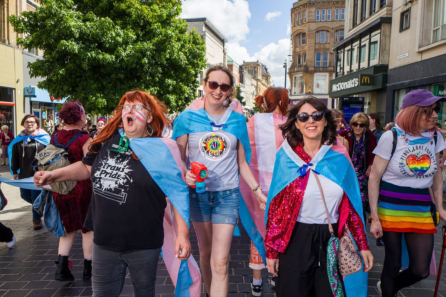 A group of trans people and allies march through Liverpool wearing rainbows and trans flags. In the middle a trans woman has a "Queer all year" t-shirt on.