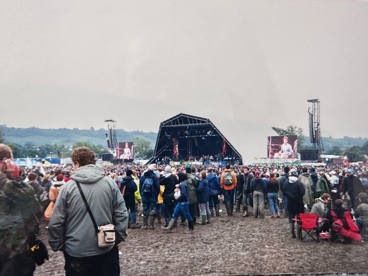 An outdoor stage with a screen at either side. Large groups of people wearing waterproof clothing stand in front. The ground is very muddy