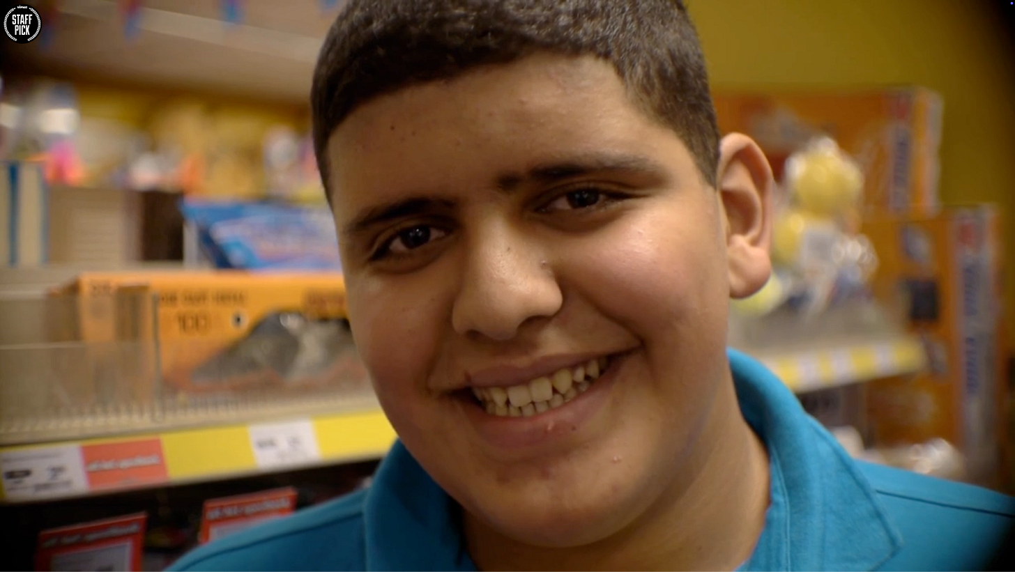 Close-up portrait of a Latino teenage boy with short dark hair wearing a bright blue polo shirt, smiling broadly at the camera in what appears to be a convenience store..