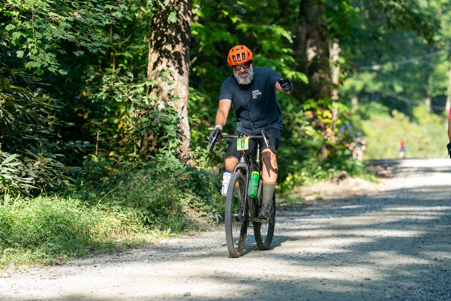 SB107036-2026-Dirt-Diggler.jpg The author rides a gravel bike on a dirt road during the 2026 Dirt Diggler race. He wears an orange helmet, dark cycling kit, and sports a full gray beard. His race number 12 is mounted on the handlebars. Lush green forest lines the road, with other riders visible in the distance.